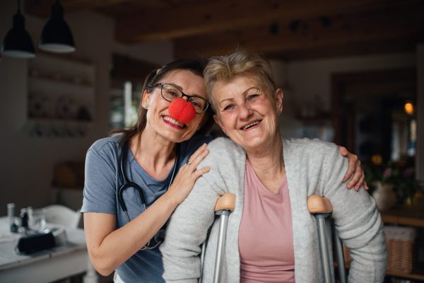 A healthcare worker or caregiver with red nose visiting senior woman indoors at home, looking at camera.